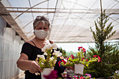 Einkäuferin mittleren Alters mit Textilmaske, die während einer Coronavirus-Pandemie in einem Gartengeschäft blühende Blumen in Töpfen auswählt