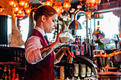 Female barkeeper pouring alcohol in shaker while preparing refreshing cocktail at counter in bar and looking away