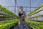Man in checkered shirt and apron checking growth of lettuce seedling and taking notes in notebook in greenhouse with hydroponic system