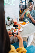 Crop anonymous female vlogger taking photo of tasty burger on cellphone against black friend with smoothie at table