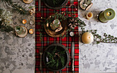 Draufsicht auf einen weihnachtlich gedeckten Tisch mit Kranz auf dem Teller, dekorativem Holzschmuck und rot-karierter Tischdecke mit gelben Lichtern im Hintergrund