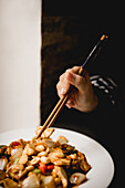 Unrecognizable female using chopsticks to eat portion of delicious Gong Bao chicken against black background in restaurant