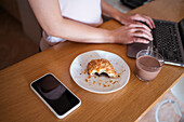 Crop anonymous female browsing modern netbook while sitting at wooden counter with pastry and hot drink near smartphone in apartment