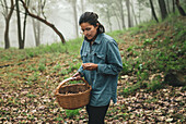 Serious female in eyeglasses carrying wicker basket and picking edible mushrooms in woods in hazy weather