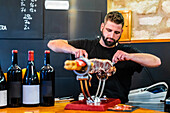 Bearded professional ethnic cutter preparing tasty jamon in restaurant with bottles of wine