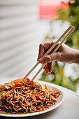 Hand isst gebratene leckere appetitliche Nudeln mit gesundem Gemüse mit Stäbchen in einem asiatischen Cafe