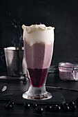 Glass of tasty smooth banana and blueberry drink with whipped cream on top on cutting board on dark background