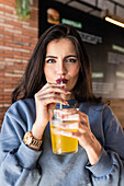 Fröhliche junge Frau im blauen Pullover nippt an einem kalten Erfrischungsgetränk durch einen Strohhalm, während sie ihre Freizeit in einer Cafeteria verbringt und fröhlich in die Kamera schaut