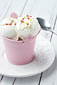 Blick von oben auf appetitliches Vanilleeis, serviert in einem kleinen rosa Metalleimer und dekoriert mit süßen sternförmigen Streuseln auf einem weißen Holztisch bei Tageslicht
