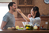Side view of cheerful young Asian female with long dark hair in casual clothes feeding happy Hispanic husband with cheese while preparing delicious salad in kitchen