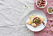 Draufsicht auf leckere Crêpes, die mit Schokolade und Nüssen garniert auf einem Teller auf dem Frühstückstisch serviert werden
