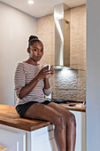 Side view of young dreamy African American female with mug of hot drink sitting on table and looking at camera indoors
