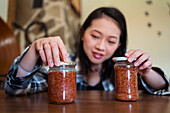 Young ethnic female with glass jars of delicious fig marmalade on table in house on blurred background