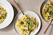 Draufsicht auf appetitlich gekochte Ravioli mit grüner Soße und Kräutern auf weißen Tellern mit Gabeln auf dem Tisch