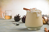 Glass of milk punch with cinnamon powder on whipped egg white against pine cones on Christmas Day on light background