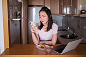 Positive asiatische Frau schreibt Textnachrichten auf dem Handy, während sie am Tisch mit Netbook und Essen mit heißem Getränk in der Küche sitzt