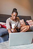 Young black female with cup of hot drink browsing internet on netbook while sitting on sofa in house room
