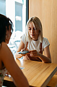 Best female friends surfing internet on cellphone while talking at cafeteria table in daylight