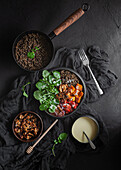 Draufsicht auf Eintopf mit Linsen neben Salat mit Kürbis und Paprika, dekoriert mit grünen Basilikumblättern auf schwarzem Hintergrund neben Salatdressing