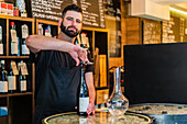 Bearded ethnic barman looking at camera and opening glass bottle with wine near vase on metal tray on counter