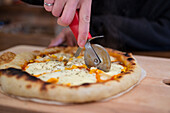 Crop female cutting tasty baked pizza with sharp cutter served on wooden cutting board at table in light kitchen