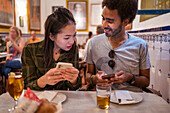 Happy young multiracial couple in casual clothes using smartphones while sitting together at table with glasses of beer in modern restaurant