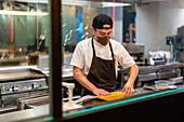 Side view of male cook in protective mask standing at stainless counter with steel tray of baked flatbread in restaurant open kitchen during pandemic