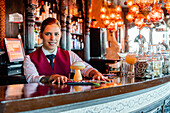 Smiling female barkeeper standing at bar counter with a type of alcohol drink served in creative cocktail glasses in shape of mushroom