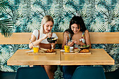 Young content best female friends sitting at wooden table with tasty croissants and beverages in cafeteria