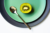From above of half of fresh ripe juicy kiwi and spoon placed on grey and green plates on white surface