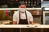 Männlicher Koch mit Schürze, weißem Hemd, Mütze und Schutzmaske bei der Zubereitung von Speisen in einem Restaurant während der Coronavirus-Krise und Blick in die Kamera