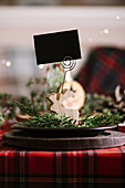 Weihnachtlich gedeckter Tisch mit Rentier aus Holz mit leerer Namenskarte auf Keramikteller auf rot-kariertem Tischtuch im Hintergrund