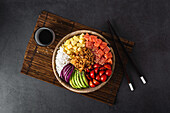 Draufsicht auf eine appetitliche bunte Poke-Schale mit frischem Lachs und Reis, serviert mit reifen Kirschtomaten, Avocadoscheiben, Zwiebeln und Mango auf einem Bambustablett mit Sojasauce
