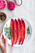 Top view of crop unrecognizable person showing gravlax with mixed peppercorns and fresh dill sprigs on plate on light background