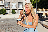 Schöne blonde junge Frau isst kaltes leckeres Eis, während sie an einem sonnigen Sommertag auf einer Straße in der Stadt sitzt