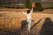 Back view of ethnic female with straw hat standing near dry hay stack in field