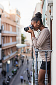 Side view ethnic female in wear with striped ornament with professional photo device looking away on balcony in daytime