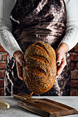 Crop anonymous female baker holding bread with sunflower seeds on table in bakehouse