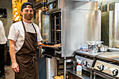 Seitenansicht eines männlichen Kochs in Uniform und Schutzmaske, der in der Küche eines Restaurants während einer Coronavirus-Pandemie ein Gericht im Ofen zubereitet
