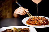 Crop female in stylish dress sitting in Asian restaurant and eating prepared tasty spicy chicken with chopsticks