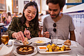 Content young Asian female millennial dipping bread into sauce of appetizing gambas al ajillo dish with shrimps while having dinner with positive ethnic boyfriend