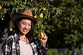 Delighted ethnic female farmer standing with pear in summer garden in countryside and looking at camera