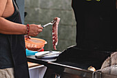 Abgeschnittene, nicht erkennbare männliche Person in Schürze, die rohes, mariniertes Fleisch auf den Grillrost eines Grillofens legt, während sie Steaks zubereitet