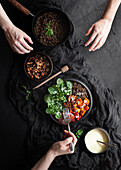 Top view of crop anonymous people at table with lentils eating salad with basil leaves and vegetables on black background