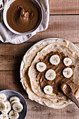 Top view of pieces of fresh banana on palatable crepes with nut butter on timber table