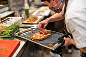 Cropped unrecognizable male cook adding filling on flatbread on stainless tray wearing protective mask and uniform in restaurant