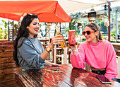 Happy young female friends in casual outfits clinking beer bottle and glass of berry cocktail while sitting at table in sunny outdoor bar