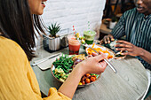 Cheerful female at table with appetizing meal and smoothies against crop anonymous ethnic partner in restaurant