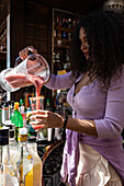 Professionelle Barkeeperin in Schürze gießt frischen Beeren-Slush-Cocktail aus dem Mixer in ein Glas, während sie in einer sonnigen Bar im Freien arbeitet