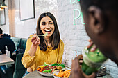 Zufriedene Frau, die ein leckeres Gericht genießt, während sie sich mit einem nicht erkennbaren ethnischen Partner unterhält, der einen Smoothie im Restaurant trinkt